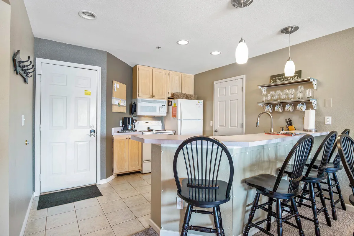This image features a spacious kitchen interior with a kitchen island, black bar stools, a white refrigerator, and kitchen cabinets. The arrangement suggests a welcoming space designed for both meal preparation and socializing, with ample counter space for cooking and serving.