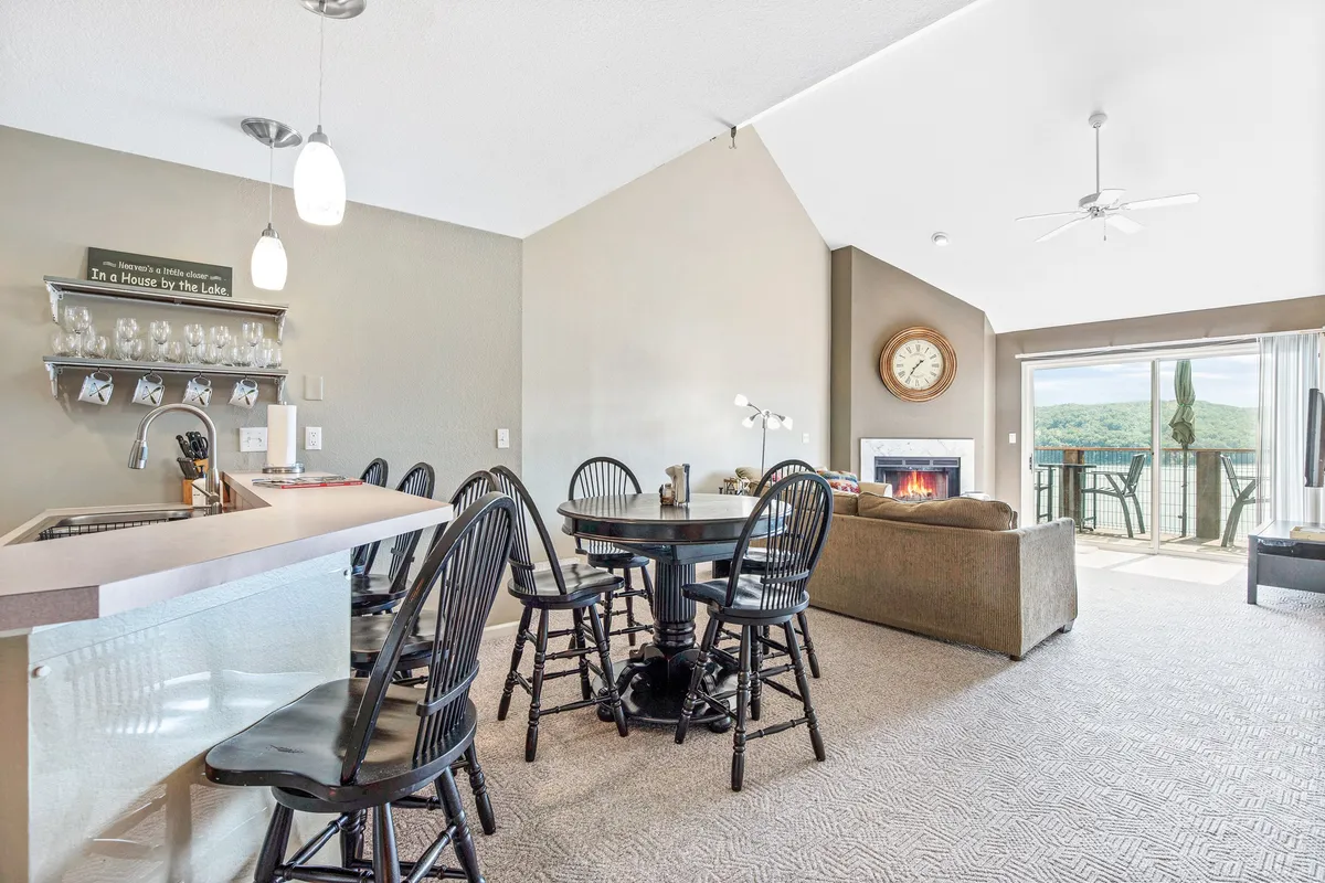 This image captures a bright, open-concept interior with a kitchen island, dining table, comfortable sofa, fireplace, and a view through sliding doors to a deck. The space is bathed in natural light, highlighting how the living, dining, and kitchen areas flow seamlessly together. The deck beyond suggests a connection to the outdoors and beautiful lake views.