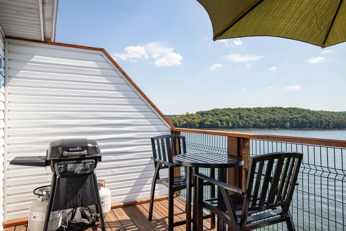 An outdoor patio with a grill, table and chairs shaded by an umbrella, and a stunning lake view in the background. The scene suggests outdoor dining and socializing in a lakeside setting, with the soothing ambiance of the water close by.