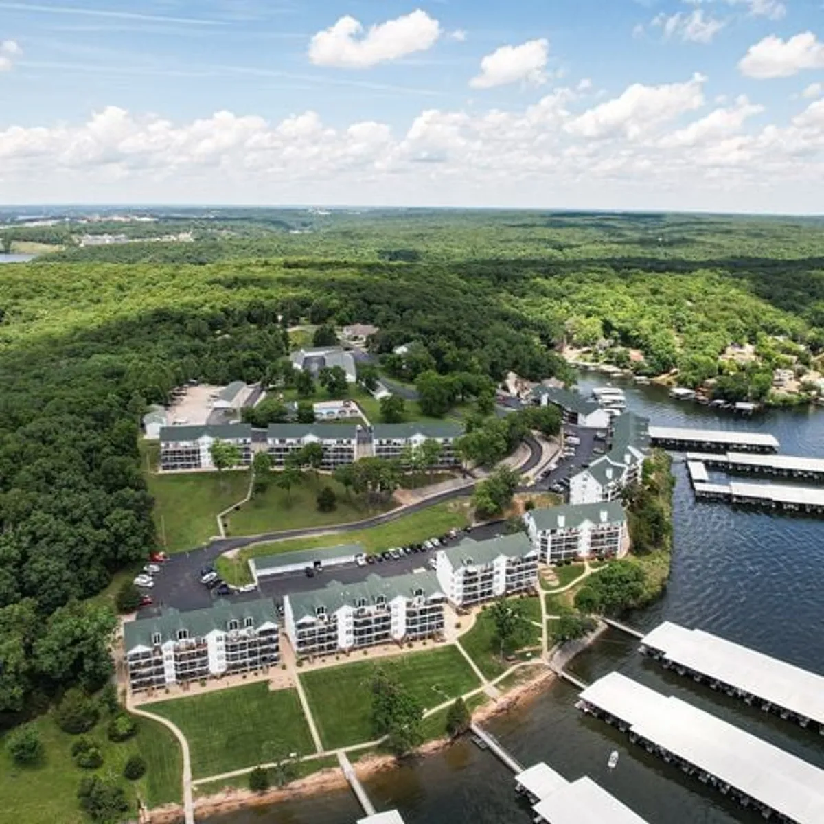 A picturesque lakeside area featuring rows of marina docks stretching out into the water, surrounded by lush green woods and multi-story residential buildings. The peaceful shoreline is ideal for boating, fishing, and picnicking, with hiking trails nearby. The scene captures the essence of waterfront leisure and outdoor family fun.