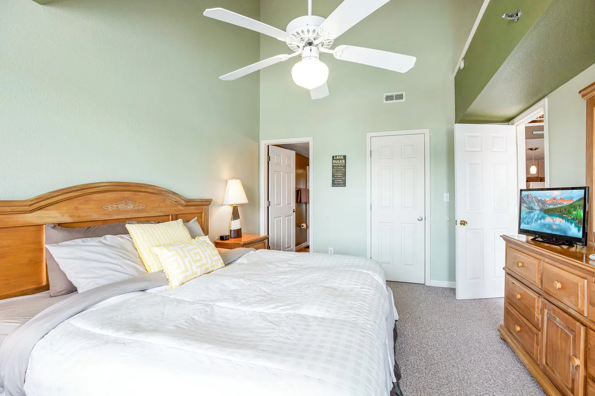 A serene, well-lit bedroom featuring a king-sized bed adorned with decorative pillows, a dresser with a TV, a ceiling fan, and a bedside lamp. Sunlight softly streams through a window, enhancing the calming atmosphere of the room.