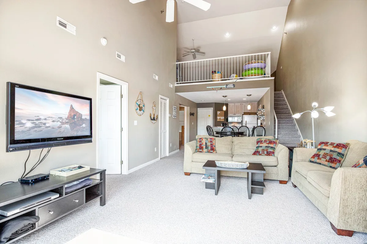 A spacious, welcoming living area featuring a cozy sofa with decorative pillows, a television, a staircase leading upstairs, and a dining area visible in the background. The layout emphasizes communal activities like movie nights, entertaining guests, and shared meals.
