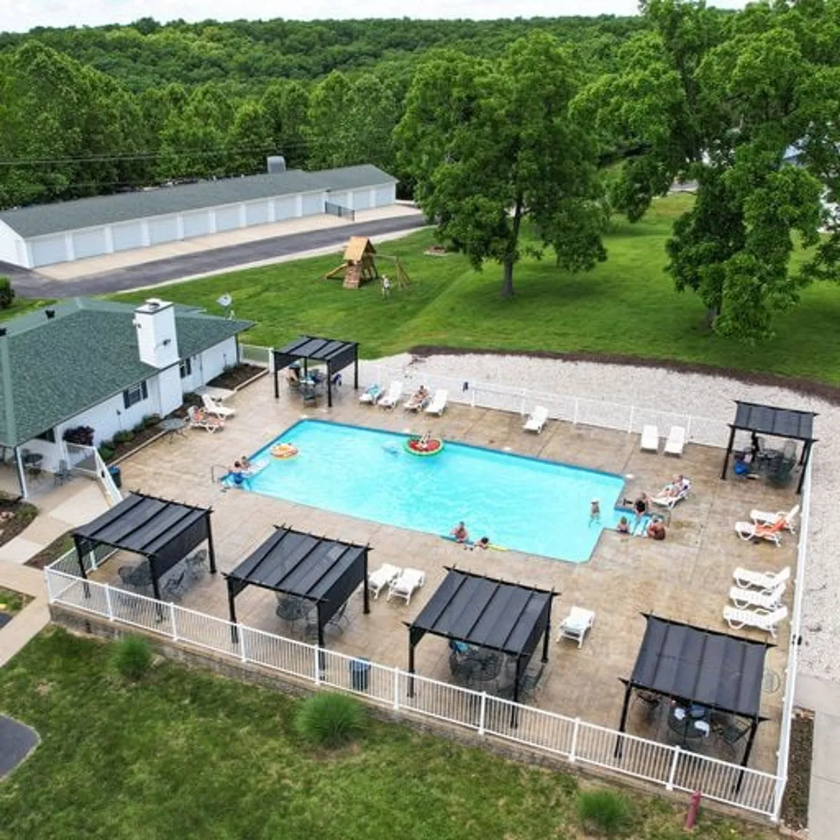 A cheerful pool area surrounded by trees, with ample seating and visible playground equipment in the background. The scene is lively and family-friendly, suggesting group activities and relaxed afternoons by the pool.