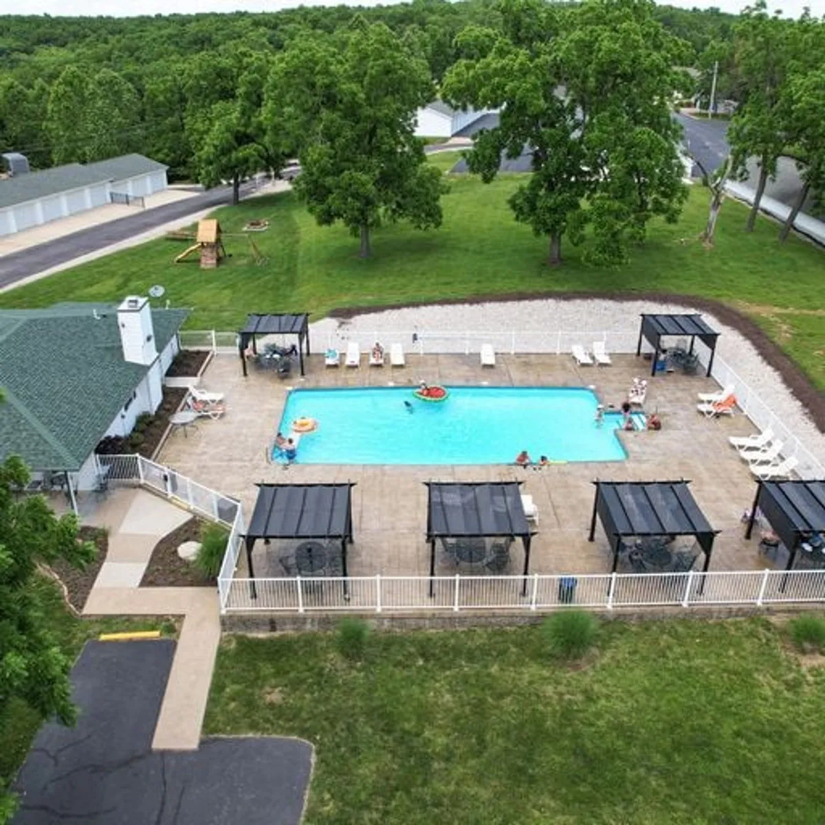 A lively outdoor swimming pool area surrounded by greenery, shade structures, and a play area for children. The scene is inviting and family-friendly, highlighting a space designed for swimming, sunbathing, and family gatherings. The pool is a cheerful hub of seasonal activity, with space for games and outdoor fun.