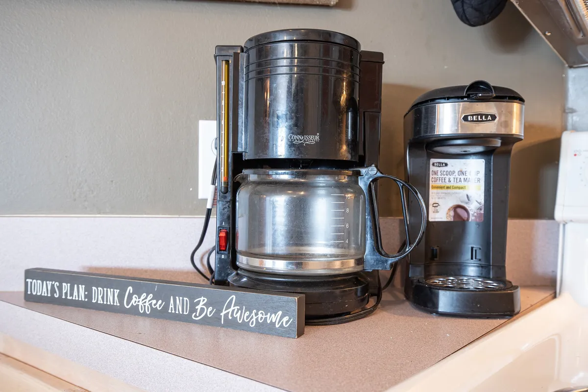 The image zooms in on a coffee maker, a single-serve coffee machine, and a decorative sign, all set up on a kitchen counter. The cheerful ambiance suggests a dedicated coffee station, ideal for morning routines or sharing a cup with guests.