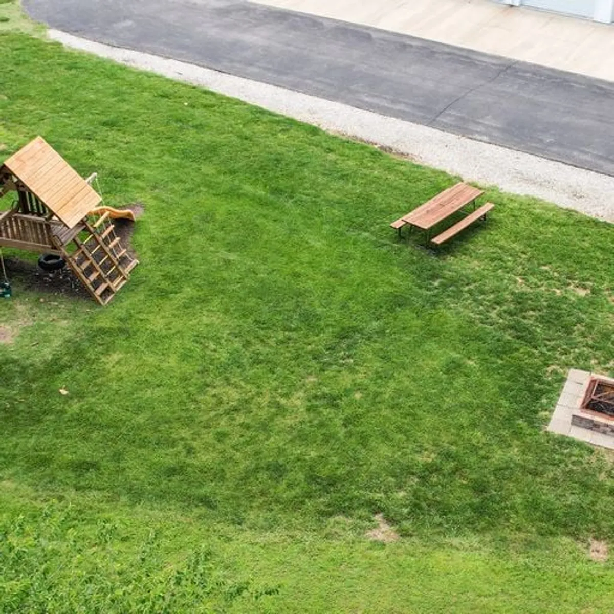 A cheerful outdoor setting featuring a playground structure, picnic table, and fire pit, all surrounded by greenery. Children play while families gather for meals or share stories around the fire. The scene is relaxed, safe, and welcoming, inviting multi-generational groups to enjoy time outdoors together.