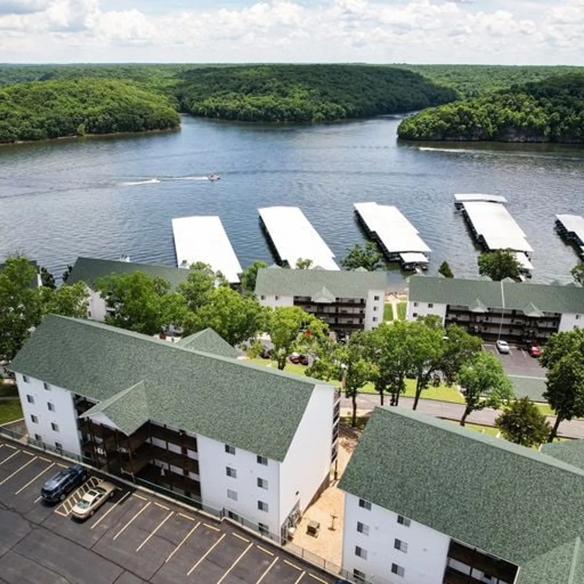 This image captures a serene lake view with a marina, residential buildings, and lush greenery. The tranquil water catches the soft light, suggesting either sunrise or sunset, and the scene is perfect for boating, fishing, and relaxing walks along the shore.