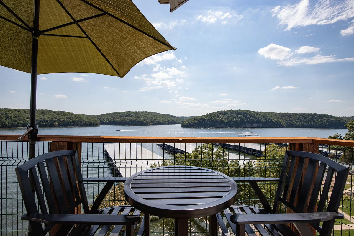 A tranquil view from a wooden table and two chairs under a green umbrella on the deck, overlooking the lake and distant hills. The scene suggests relaxation, conversation, and shared moments with a scenic backdrop.